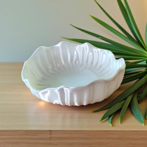 May include: A white, scalloped-edge ceramic bowl with a textured interior. The bowl is pearlescent and sits on a light-colored wooden surface. Green plant leaves are visible in the background, adding a natural element to the composition.