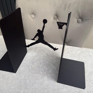 May include: Black metal bookends featuring a basketball player silhouette mid-air, reaching for a basketball. One bookend is a solid panel, the other has a hoop. A decorative item for a sports fan.