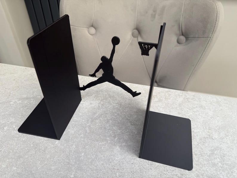 May include: Black metal bookends featuring a basketball player silhouette mid-air, reaching for a basketball. One bookend is a solid panel, the other has a hoop. A decorative item for a sports fan.