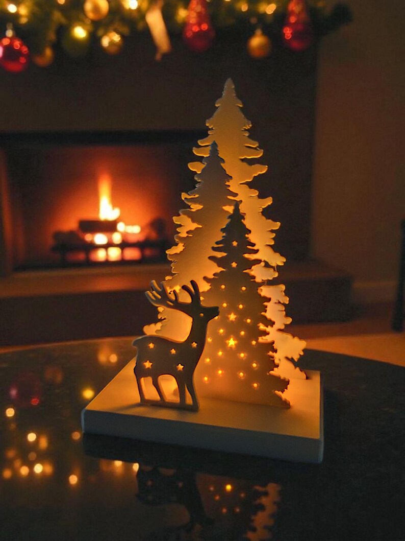 May include: A decorative Christmas scene featuring a wooden reindeer and two Christmas trees with star-shaped cutouts. The trees and reindeer are illuminated with warm, soft light. The scene is set on a table with a fireplace in the background.
