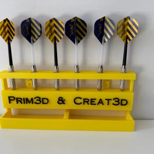 Puede incluir: Un soporte amarillo para dardos impreso en 3D con seis dardos. Los dardos tienen ca&ntilde;as negras y plateadas y plumas a rayas amarillas y azul marino. El soporte tiene el texto "PRIM3D & CREAT3D" en negro.