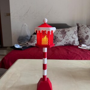 Op de afbeelding: Een rood-witte decoratieve lantaarn met een nepkaars erin. De lantaarn heeft een rood dak met witte rand en een witte bal bovenop. De paal is rood-wit gestreept en de basis is rood.
