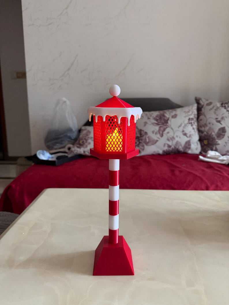 Op de afbeelding: Een rood-witte decoratieve lantaarn met een nepkaars erin. De lantaarn heeft een rood dak met witte rand en een witte bal bovenop. De paal is rood-wit gestreept en de basis is rood.