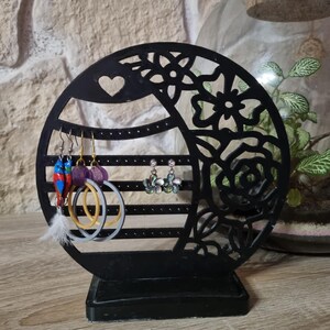 May include: A black, laser-cut earring holder with a circular design featuring floral and heart cutouts. The holder displays various earrings, including hoops and beaded styles. The base is rectangular, and the background includes a stone wall and a terrarium.