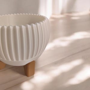 Pode incluir: Um vaso de cer&acirc;mica branco, canelado, com uma borda ondulada, apoiado em tr&ecirc;s pequenas pernas castanho-claro. O vaso tem uma forma arredondada e &eacute; projetado para uso interno. O fundo &eacute; uma superf&iacute;cie de madeira clara.