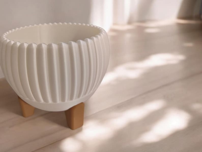 Pode incluir: Um vaso de cer&acirc;mica branco, canelado, com uma borda ondulada, apoiado em tr&ecirc;s pequenas pernas castanho-claro. O vaso tem uma forma arredondada e &eacute; projetado para uso interno. O fundo &eacute; uma superf&iacute;cie de madeira clara.