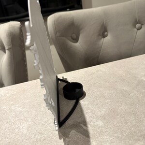 Pu&ograve; includere: Un portacandele decorativo bianco con base in metallo nero. Il portacandele ha un design dettagliato e uno spazio circolare per una candela. Il supporto &egrave; su una superficie grigio chiaro.