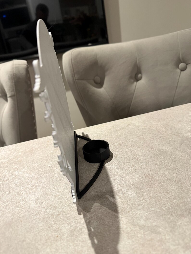 Pu&ograve; includere: Un portacandele decorativo bianco con base in metallo nero. Il portacandele ha un design dettagliato e uno spazio circolare per una candela. Il supporto &egrave; su una superficie grigio chiaro.
