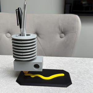 May include: A gray and black pen holder with a unique design. The pen holder has a cylindrical top section with a series of horizontal ridges, a rectangular base, and a black base with a yellow line. Two pens are in the holder.