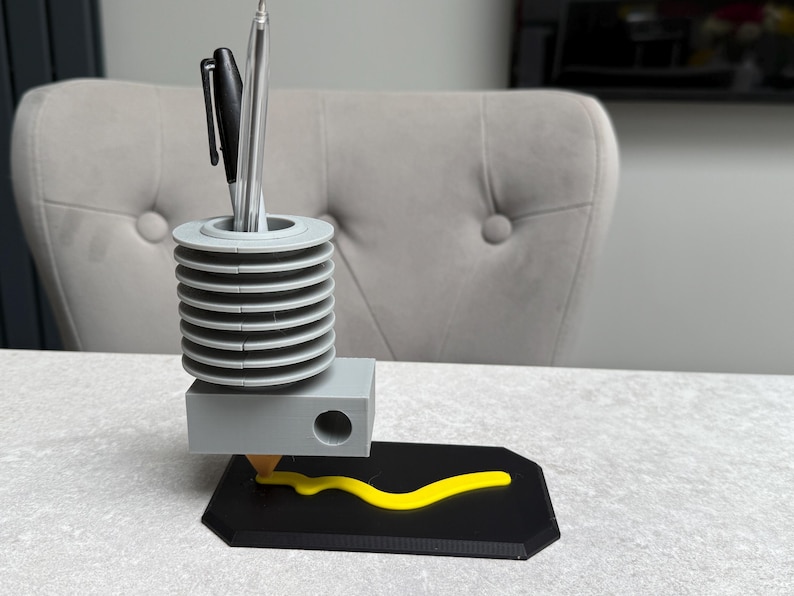 May include: A gray and black pen holder with a unique design. The pen holder has a cylindrical top section with a series of horizontal ridges, a rectangular base, and a black base with a yellow line. Two pens are in the holder.