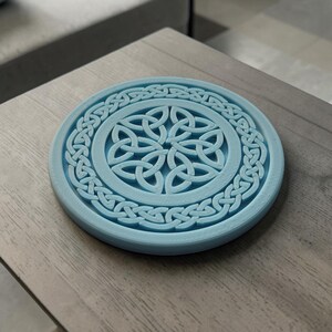 May include: A round, light blue coaster with a Celtic knot design. The coaster features a central knot pattern surrounded by a braided border. The coaster is made of a solid material and sits on a wooden surface.
