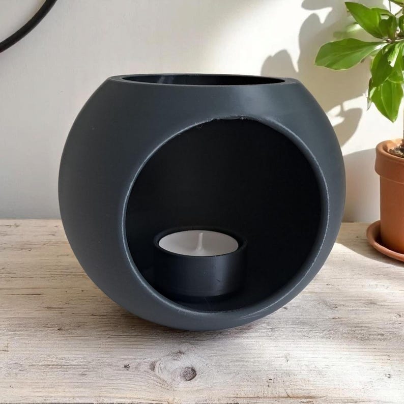 May include: A dark grey, spherical oil diffuser with a large circular opening. A lit tea light candle sits inside the diffuser. The diffuser is on a light-coloured wooden surface.