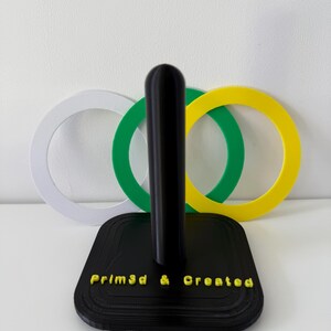 May include: A black paper towel holder with a square base and a central pole. The base has the text "Prim3d & Created" in yellow. Behind the holder are three interlocking rings in white, green, and yellow.