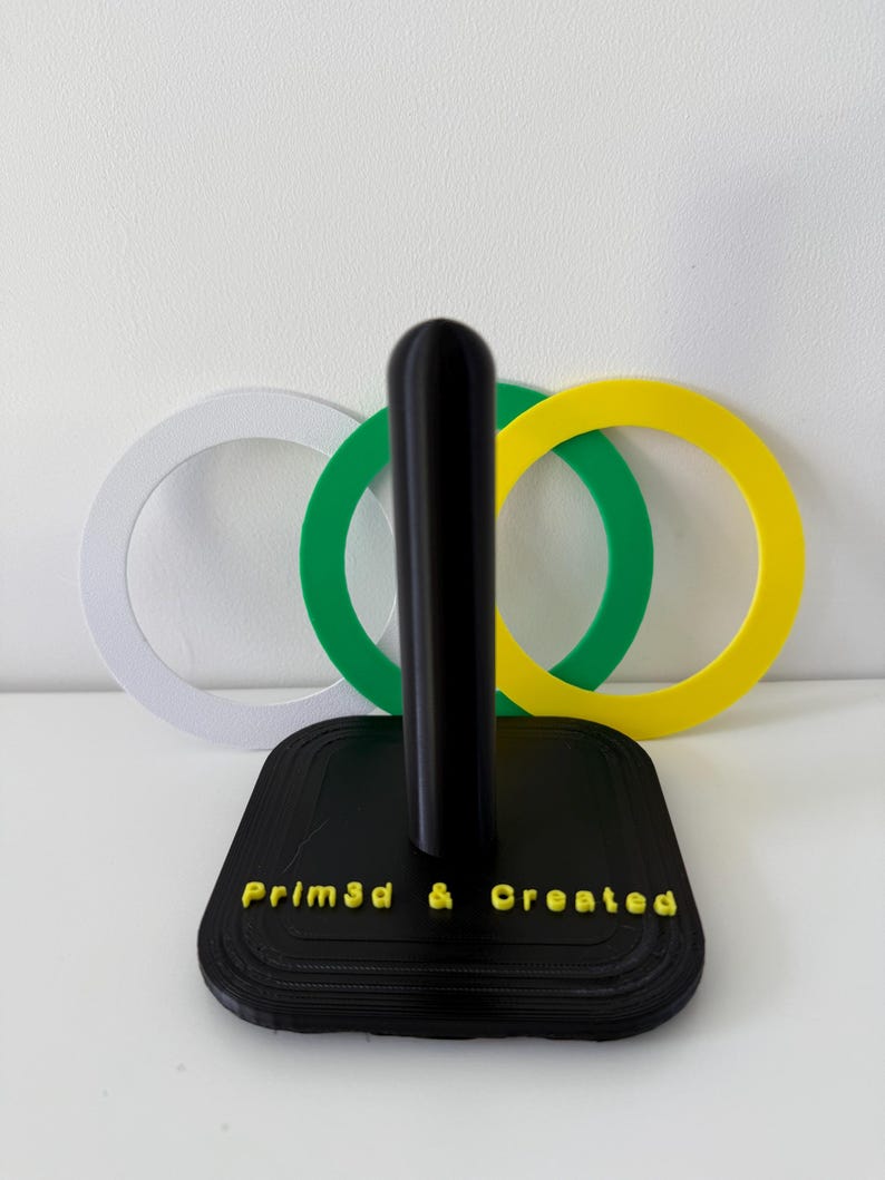 May include: A black paper towel holder with a square base and a central pole. The base has the text "Prim3d & Created" in yellow. Behind the holder are three interlocking rings in white, green, and yellow.