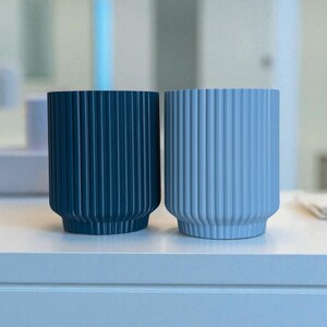 May include: Two cylindrical planters with vertical ribbed detailing. One is a deep teal color, and the other is a light blue. Both have a rounded base and are displayed on a white surface.