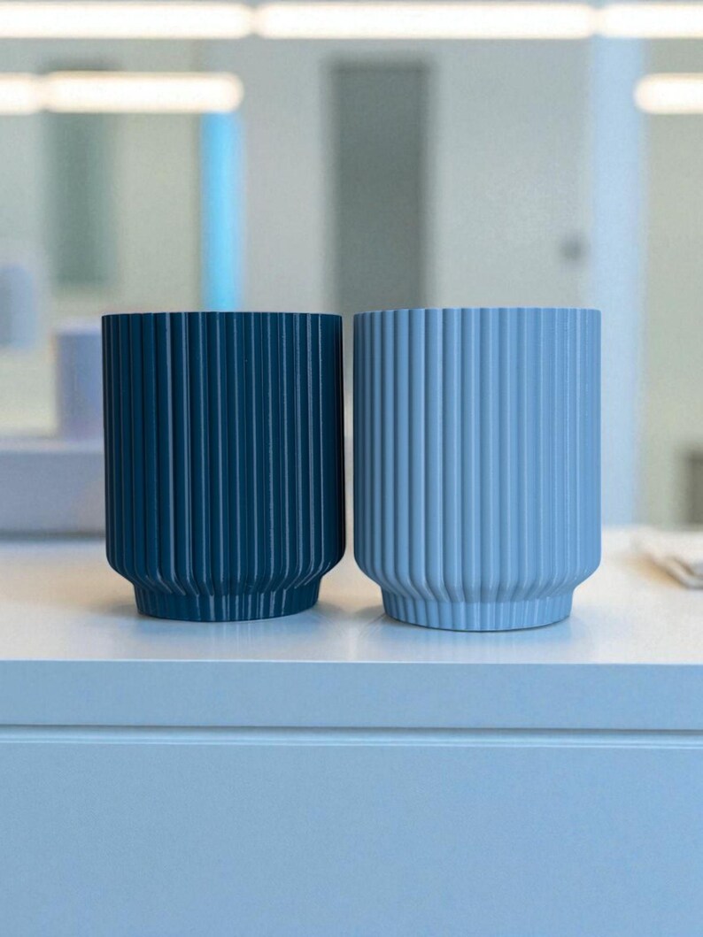 May include: Two cylindrical planters with vertical ribbed detailing. One is a deep teal color, and the other is a light blue. Both have a rounded base and are displayed on a white surface.