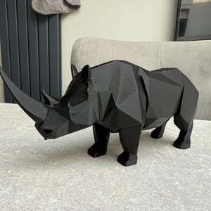 May include: A black, geometric rhino sculpture with a prominent horn. The rhino is made of a series of flat, angled planes, giving it a modern, angular appearance. The sculpture is displayed on a light-colored surface.