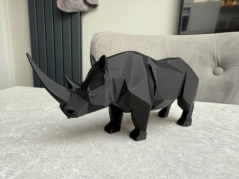 May include: A black, geometric rhino sculpture with a prominent horn. The rhino is made of a series of flat, angled planes, giving it a modern, angular appearance. The sculpture is displayed on a light-colored surface.