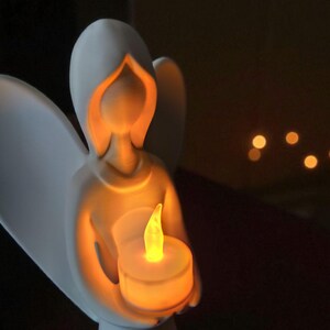 May include: A white ceramic angel figurine holding a glowing LED candle. The angel's form is simple, with a hooded head and outstretched wings. The candle emits a warm, yellow light, creating a soft glow. The background is dark, emphasizing the angel and candle.