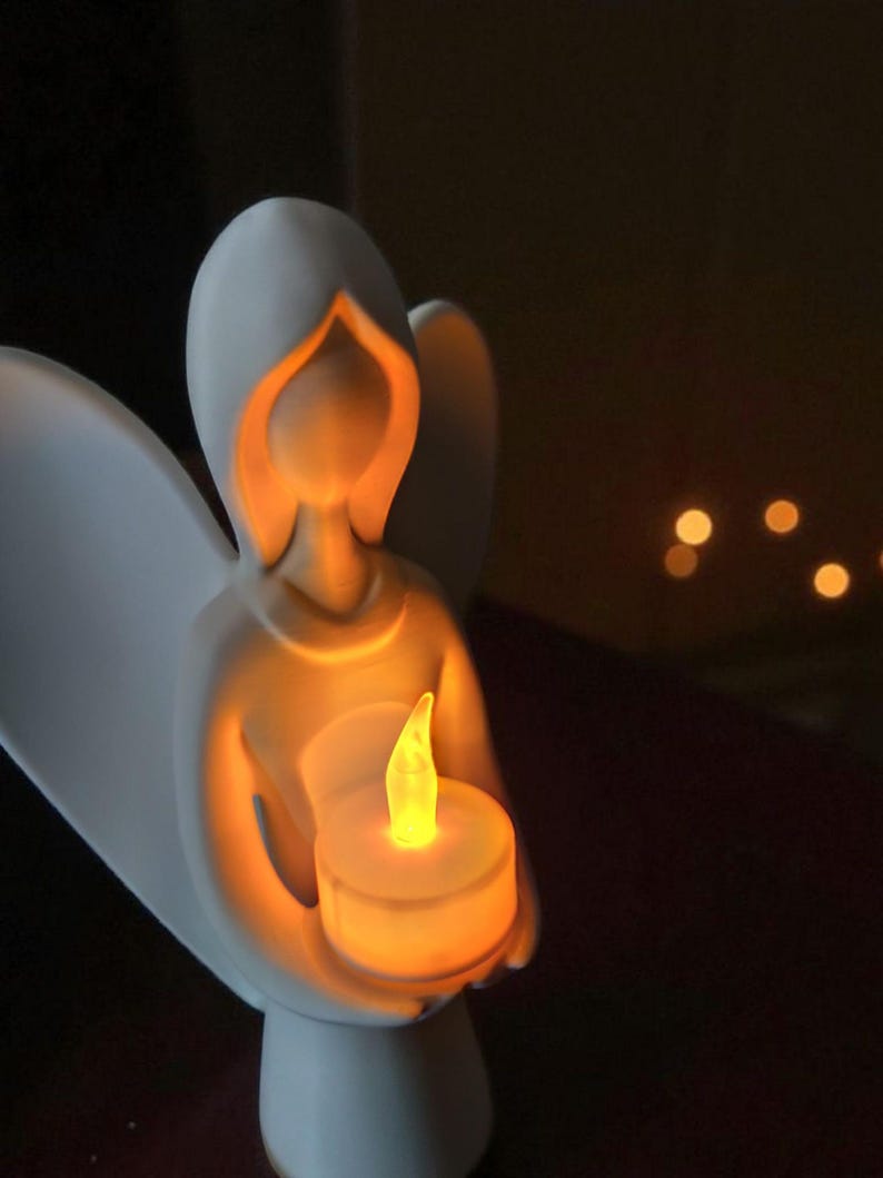 May include: A white ceramic angel figurine holding a glowing LED candle. The angel's form is simple, with a hooded head and outstretched wings. The candle emits a warm, yellow light, creating a soft glow. The background is dark, emphasizing the angel and candle.