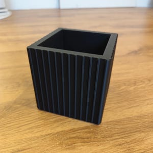 May include: A black, square planter with vertical ribbed detailing. The planter is made of a solid material and sits on a wooden surface. The design is modern and minimalist, suitable for various plants.