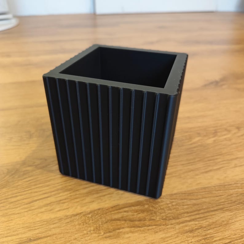 May include: A black, square planter with vertical ribbed detailing. The planter is made of a solid material and sits on a wooden surface. The design is modern and minimalist, suitable for various plants.