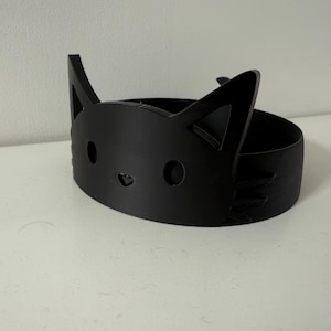 Puede incluir: Un recipiente negro con forma de gato impreso en 3D. El recipiente tiene un cuerpo circular con un dise&ntilde;o de cara de gato en la parte delantera, que incluye recortes para los ojos y la nariz, y orejas puntiagudas. El recipiente est&aacute; sobre una superficie blanca.