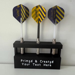 May include: Three darts with yellow and navy blue striped flights are displayed in a black holder. The flights have the text "The Nuke". The holder has the text "Prim3d & Created Your Text Here".