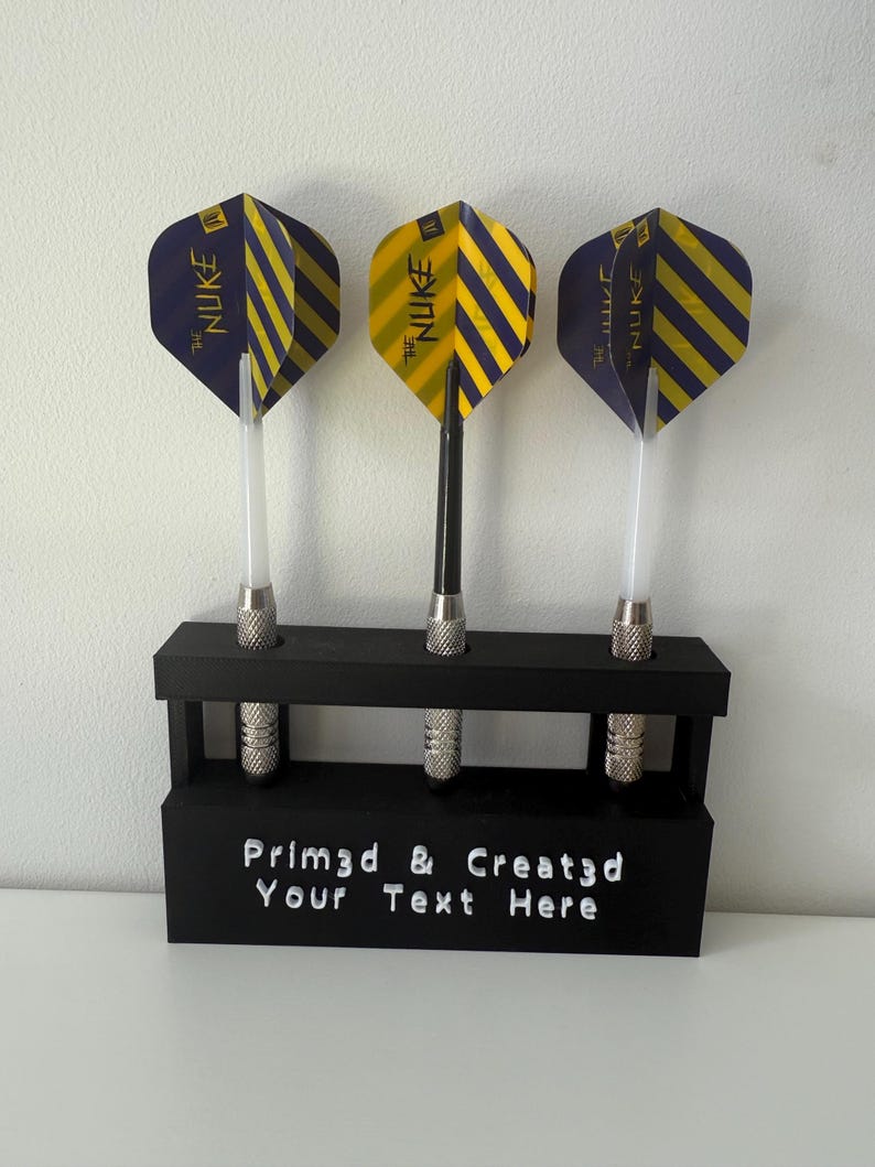 May include: Three darts with yellow and navy blue striped flights are displayed in a black holder. The flights have the text "The Nuke". The holder has the text "Prim3d & Created Your Text Here".