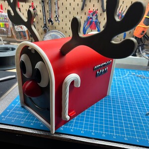 May include: A red and white Christmas mailbox with a reindeer face design. The mailbox has large cartoon eyes, a red nose, and black antlers. The words "CHRISTMAS MAIL" are on the side, and a candy cane handle is on the front.