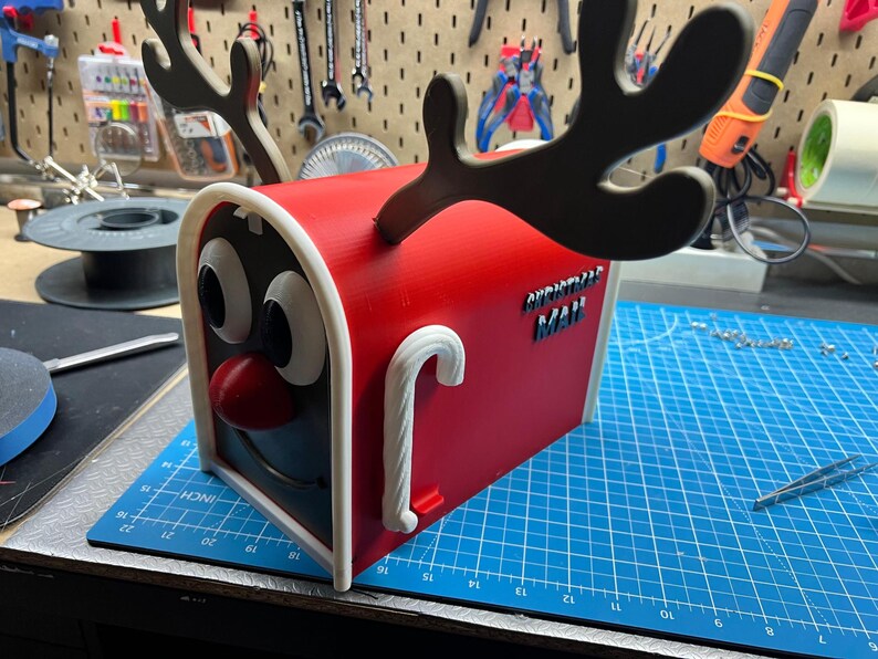 May include: A red and white Christmas mailbox with a reindeer face design. The mailbox has large cartoon eyes, a red nose, and black antlers. The words "CHRISTMAS MAIL" are on the side, and a candy cane handle is on the front.