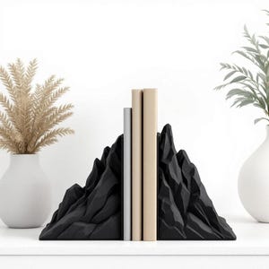 May include: Black mountain-shaped bookends with a textured surface, holding three books. The bookends are on a white surface, with vases of plants on either side. The bookends add a decorative touch to any bookshelf.
