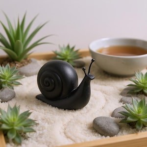 May include: A black snail figurine sits in a miniature zen garden with succulents, small stones, and a bowl of tea. The snail has a detailed shell and antennae, creating a calming, natural aesthetic.