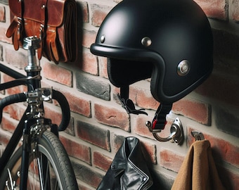 May include: A black motorcycle helmet with a rounded design, suspended on a wall. The helmet features a chin strap and is mounted on a silver hook. A black leather jacket and a brown leather bag are also visible.
