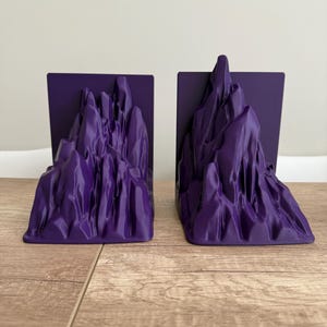 May include: A pair of purple bookends with a mountain range design. Each bookend features a rectangular base with a sculpted mountain peak. The bookends are made of a smooth, solid material and are designed to hold books upright.