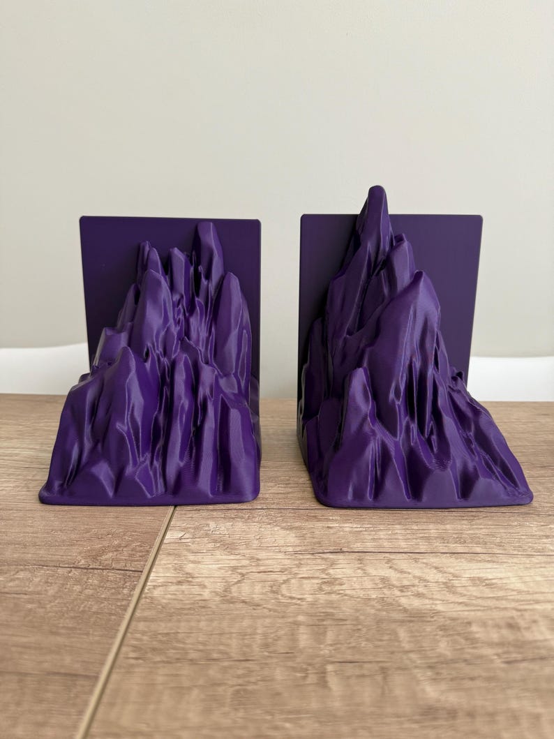 May include: A pair of purple bookends with a mountain range design. Each bookend features a rectangular base with a sculpted mountain peak. The bookends are made of a smooth, solid material and are designed to hold books upright.