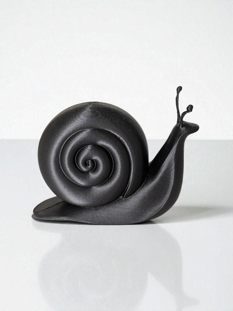 May include: A black snail figurine with a spiraled shell and two antennae. The snail is depicted in a smooth, matte finish, and is set against a white background. The figurine is a decorative item.