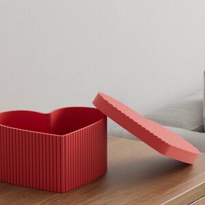 Pu&ograve; includere: Una scatola rossa a forma di cuore con un coperchio abbinato, entrambi con dettagli a coste verticali. Il coperchio &egrave; leggermente socchiuso, rivelando l'interno della scatola. La scatola &egrave; su una superficie di legno.