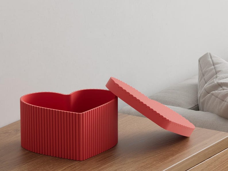 Pu&ograve; includere: Una scatola rossa a forma di cuore con un coperchio abbinato, entrambi con dettagli a coste verticali. Il coperchio &egrave; leggermente socchiuso, rivelando l'interno della scatola. La scatola &egrave; su una superficie di legno.