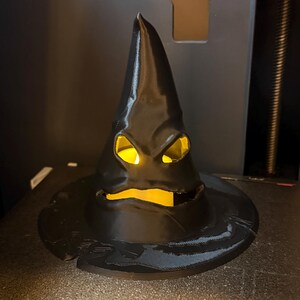 Op de afbeelding: Een zwarte, puntige hoed met een brede rand, met uitgesneden ogen en mond, verlicht door een warm, geel licht. De hoed heeft een getextureerd oppervlak en een enigszins dreigende uitdrukking, perfect voor Halloween of thema-decoratie.