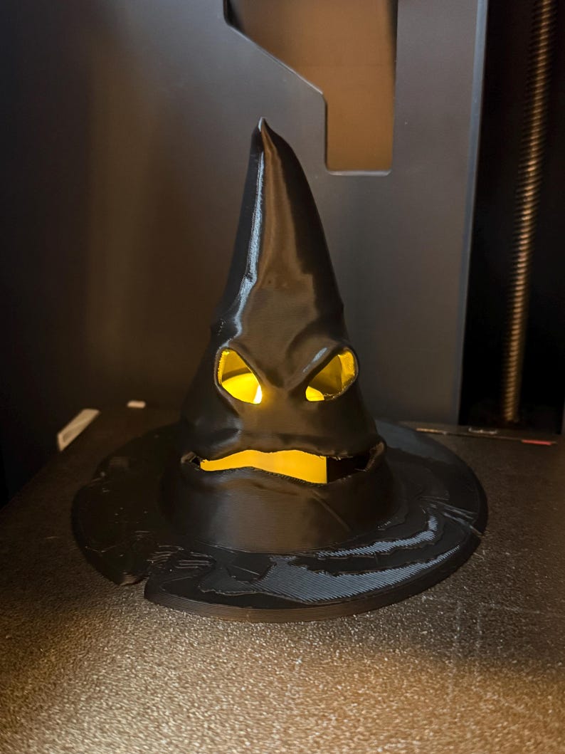 Op de afbeelding: Een zwarte, puntige hoed met een brede rand, met uitgesneden ogen en mond, verlicht door een warm, geel licht. De hoed heeft een getextureerd oppervlak en een enigszins dreigende uitdrukking, perfect voor Halloween of thema-decoratie.
