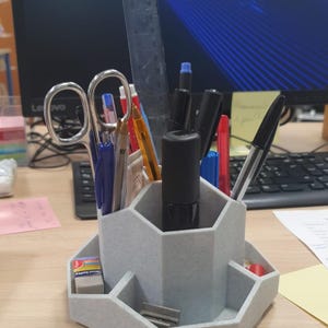 May include: A gray desk organizer with multiple compartments holding pens, pencils, scissors, and a ruler. The organizer also has a small tray with an eraser and a pencil sharpener. The background includes a computer monitor and keyboard.