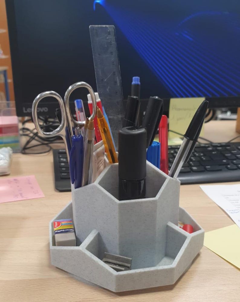 May include: A gray desk organizer with multiple compartments holding pens, pencils, scissors, and a ruler. The organizer also has a small tray with an eraser and a pencil sharpener. The background includes a computer monitor and keyboard.