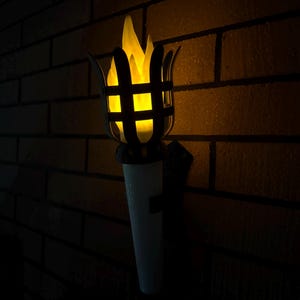 May include: An outdoor torch with a bright yellow flame effect. The torch features a black metal cage design around the flame and a white base. It is mounted on a brick wall, creating a warm ambiance.