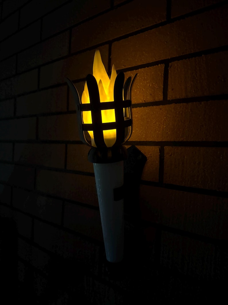 May include: An outdoor torch with a bright yellow flame effect. The torch features a black metal cage design around the flame and a white base. It is mounted on a brick wall, creating a warm ambiance.