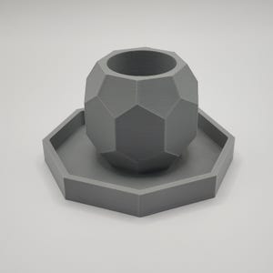 May include: A gray, geometric planter with a matching hexagonal saucer. The planter has a faceted design, and the set is made of a solid, matte material. The planter is cylindrical with a wide opening.