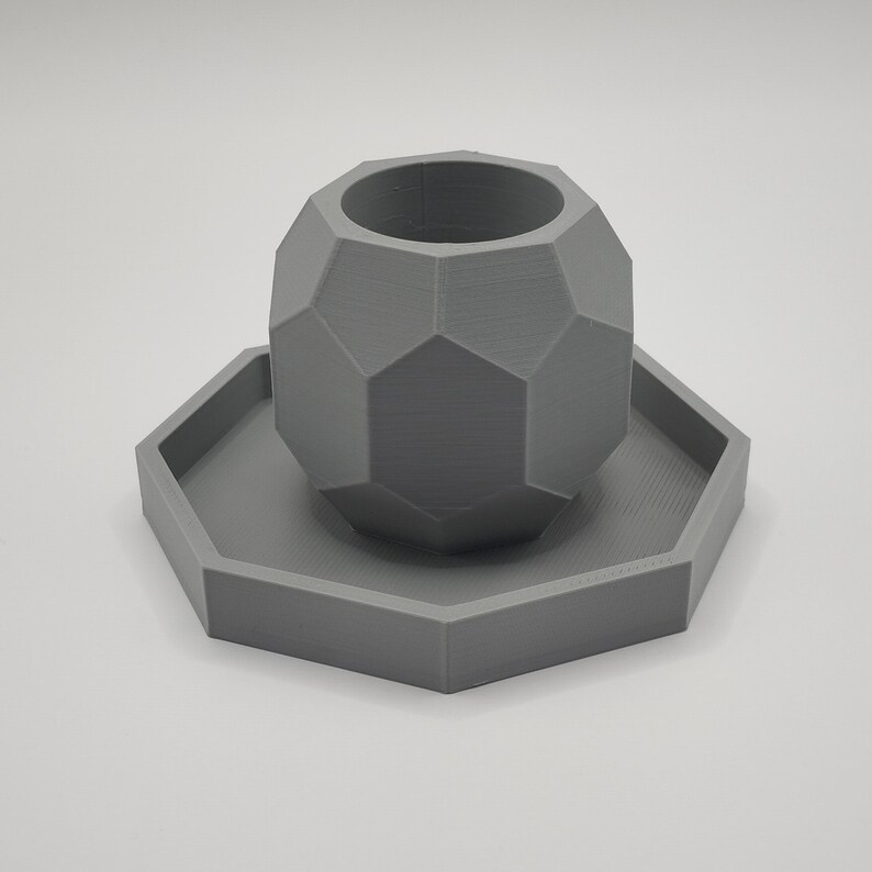 May include: A gray, geometric planter with a matching hexagonal saucer. The planter has a faceted design, and the set is made of a solid, matte material. The planter is cylindrical with a wide opening.
