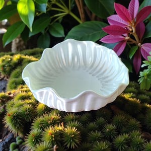 Puede incluir: Un cuenco de cer&aacute;mica blanco, con bordes festoneados y acabado nacarado. El cuenco tiene un dise&ntilde;o interior acanalado y se asienta sobre un lecho de musgo verde. El fondo presenta follaje verde y morado.