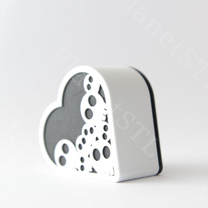 May include: A white heart-shaped box with a decorative cut-out design on the left side. The interior is dark gray. The box has a rounded shape and a black trim. The design features circles and other shapes.