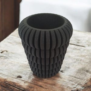 May include: A black, textured, cylindrical vase with a ribbed design. The vase has a wide opening and sits on a wooden surface. The dark color and unique texture give it a modern, minimalist aesthetic. Perfect for displaying flowers or as a decorative piece.